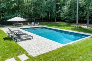 Rectangular swimming pool surrounded by a stone patio, with lounge chairs and a striped umbrella nearby, set against a backdrop of lush green trees. - pool size & what is a good size for a swimming pool - pool thermometer - pool pollen removal tips