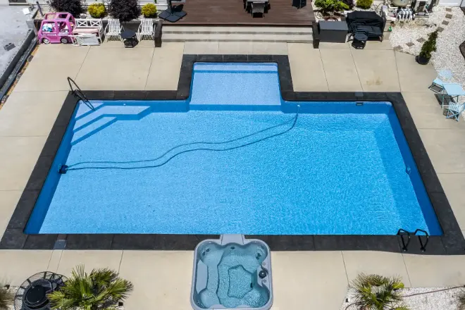 Aerial view of a rectangular swimming pool with a connected hot tub in a backyard. Pool cleaning equipment is visible inside the water. A pink toy car is parked nearby. - Prestige Pools Aerial view of a rectangular swimming pool with a connected hot tub in a backyard. Pool cleaning equipment is visible inside the water. A pink toy car is parked nearby. A vinyl liner pool with a large tanning ledge